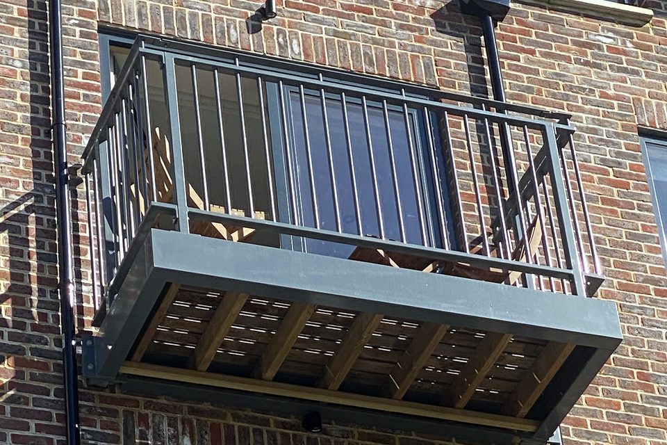 Grey steel balcony with a slatted floor and upright bar railings on a red-brick block of flats