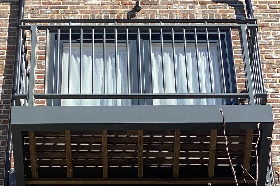 Underside of a grey steel balcony showing its slatted floor and bar railings on a brick block of flats